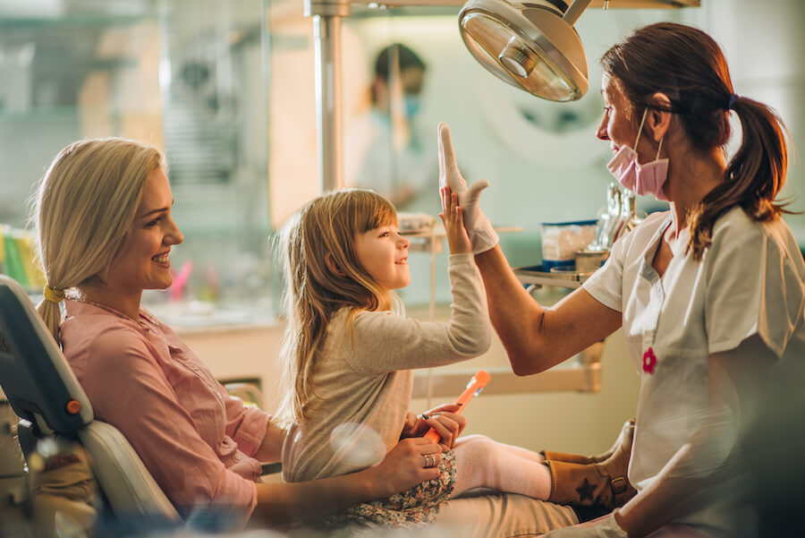 Brunette girl sitting on her mom's lap in the dental chair smiles as she high-fives her dentist