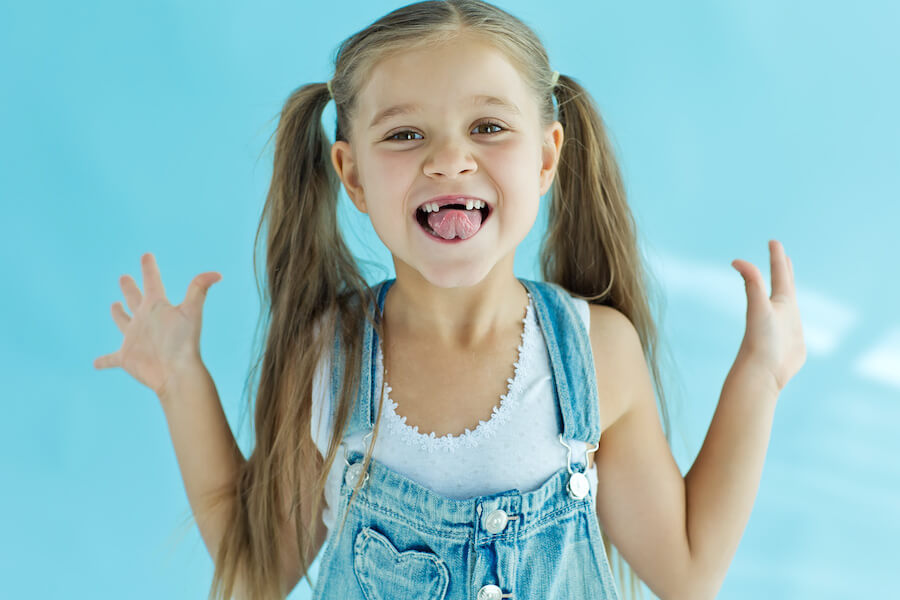 Blonde little girl with pigtails and overalls smiles to show her missing front teeth
